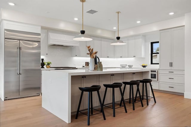 kitchen featuring decorative light fixtures, a kitchen island with sink, stainless steel appliances, light countertops, and premium range hood