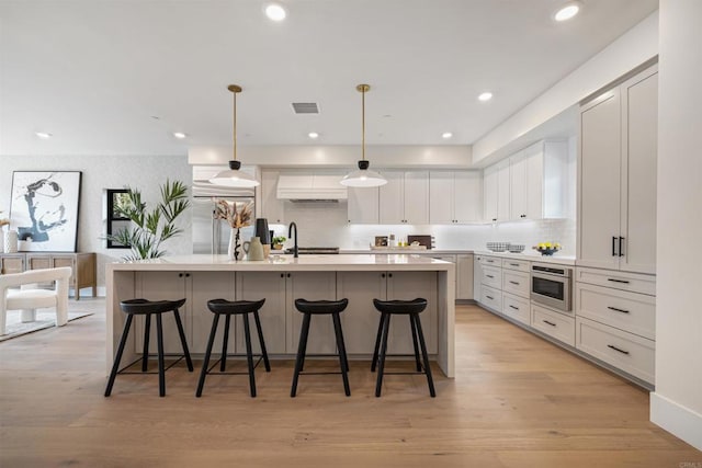 kitchen featuring a center island with sink, white cabinets, stainless steel microwave, hanging light fixtures, and light countertops