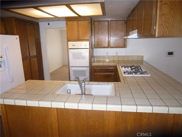 kitchen featuring tile countertops, a peninsula, white appliances, a sink, and brown cabinets