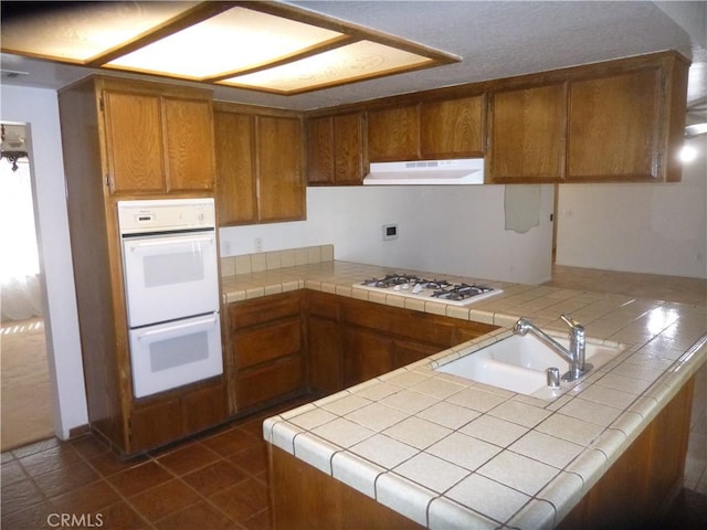 kitchen with a peninsula, white appliances, a sink, range hood, and brown cabinetry