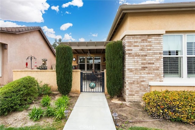 doorway to property featuring a ceiling fan, stucco siding, a gate, fence, and brick siding