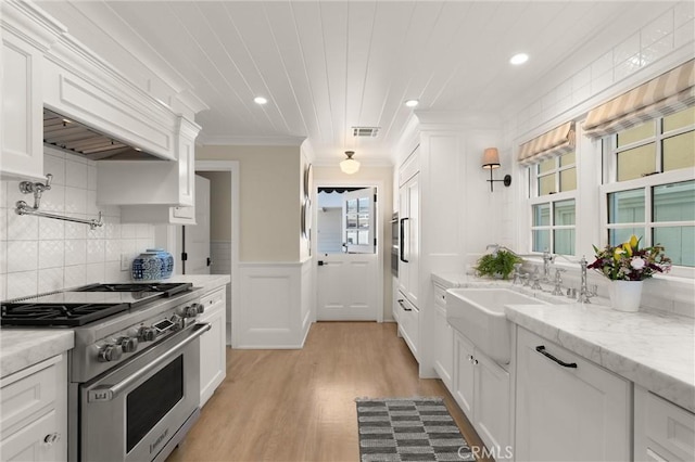 kitchen with light stone counters, high end range, ornamental molding, white cabinetry, and a sink