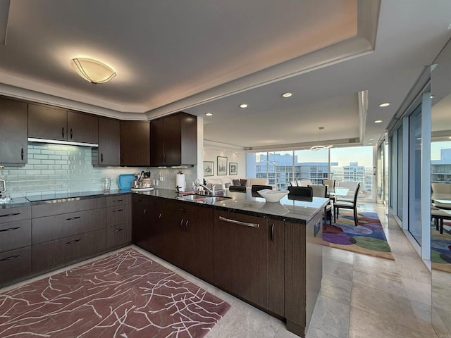 kitchen featuring a peninsula, a tray ceiling, a sink, dark brown cabinets, and backsplash