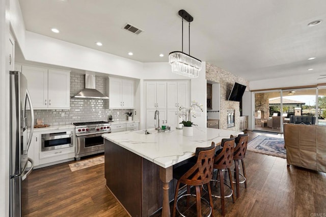 kitchen with white cabinetry, open floor plan, wall chimney range hood, appliances with stainless steel finishes, and a center island with sink