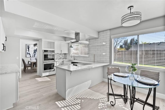 kitchen with double oven, white cabinetry, a sink, island range hood, and a peninsula