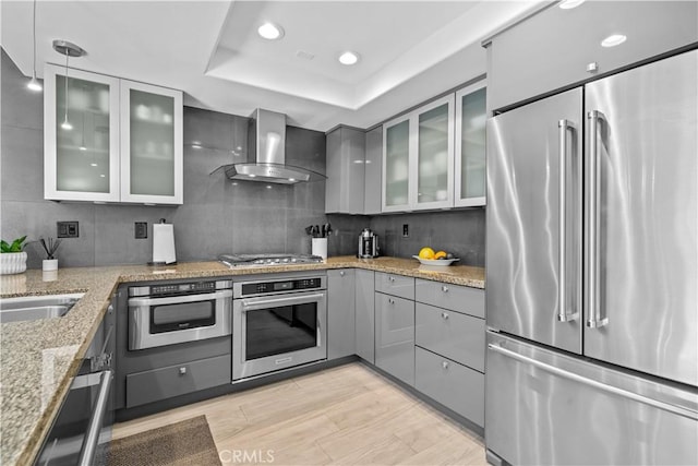 kitchen featuring a raised ceiling, wall chimney exhaust hood, gray cabinets, stainless steel appliances, and backsplash