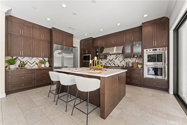 kitchen with built in appliances, extractor fan, glass insert cabinets, light countertops, and a center island with sink