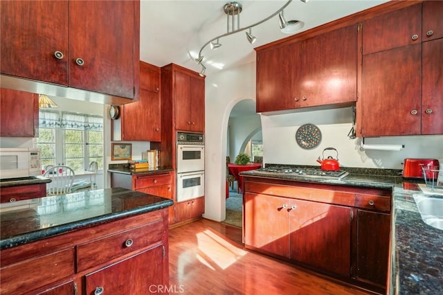 kitchen featuring arched walkways, white appliances, dark stone countertops, and light wood-style floors