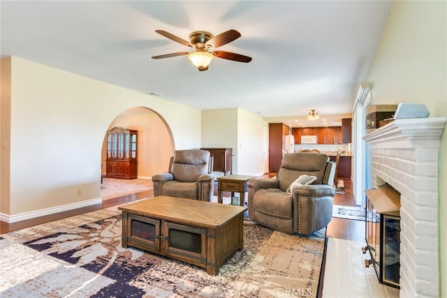 living area featuring arched walkways, a fireplace, a ceiling fan, light wood-type flooring, and baseboards