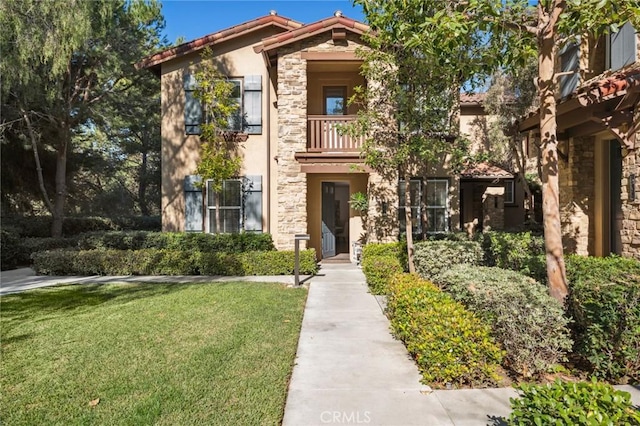 view of front of home with stucco siding, a front yard, a balcony, stone siding, and a tiled roof