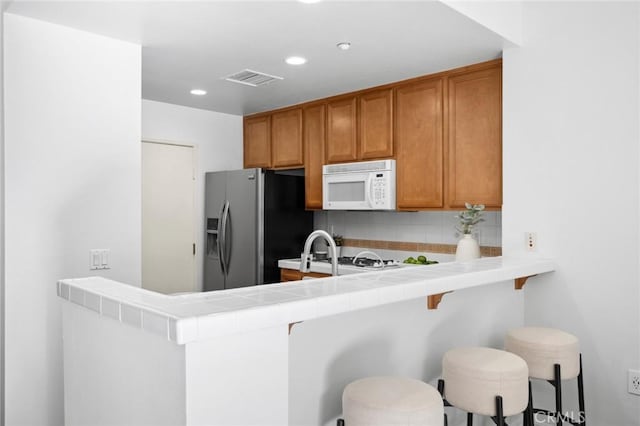 kitchen with tile countertops, white microwave, a peninsula, a kitchen breakfast bar, and stainless steel fridge