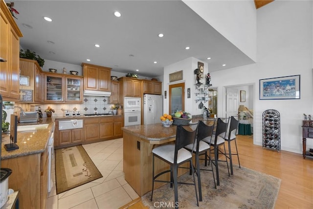 kitchen with light stone counters, a breakfast bar area, white appliances, a center island, and glass insert cabinets