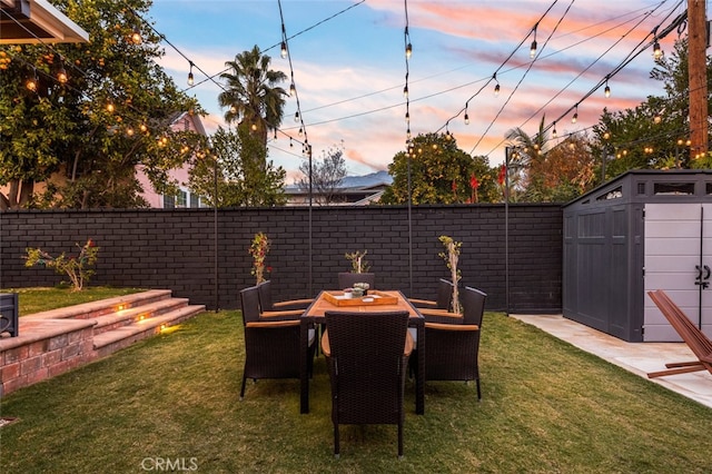 patio terrace at dusk featuring an outbuilding, a fenced backyard, a lawn, and a storage unit