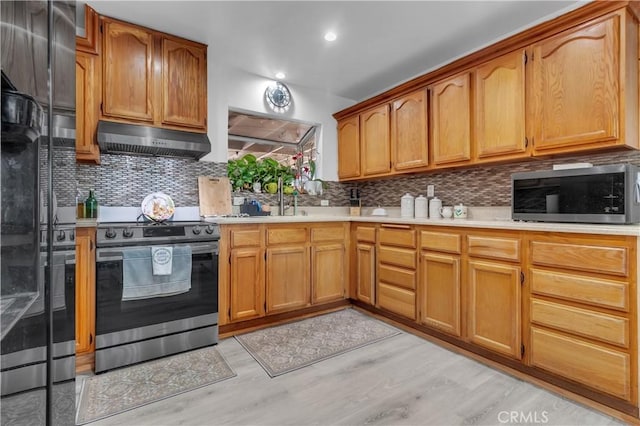 kitchen featuring tasteful backsplash, light countertops, appliances with stainless steel finishes, light wood-type flooring, and under cabinet range hood