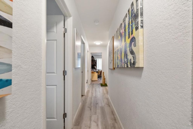 corridor featuring a textured wall, light wood-style flooring, and baseboards