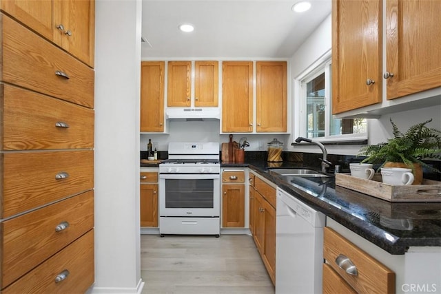 kitchen with under cabinet range hood, white appliances, a sink, brown cabinetry, and dark stone countertops