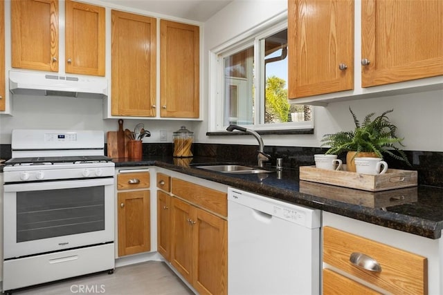 kitchen with under cabinet range hood, white appliances, a sink, brown cabinets, and dark stone countertops