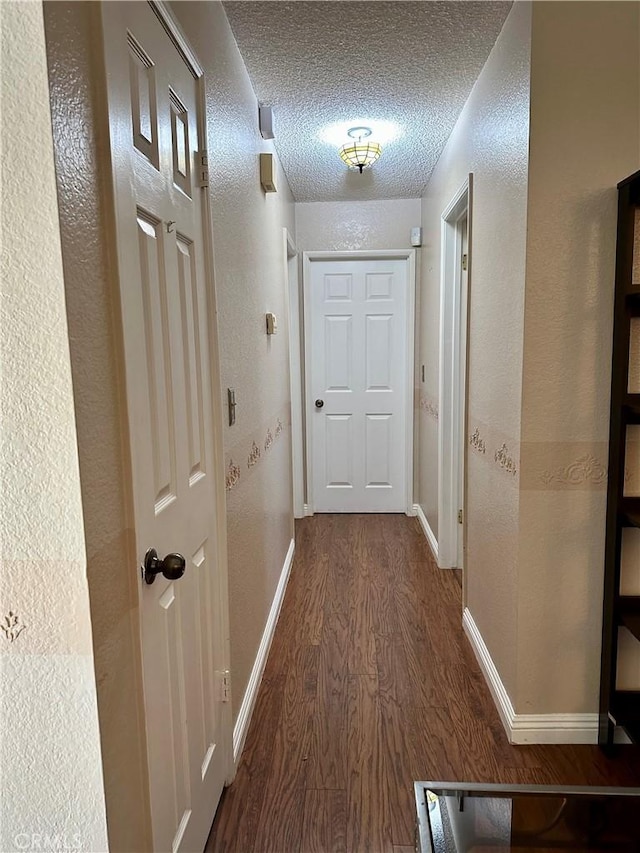 hall with dark wood-type flooring, a textured wall, a textured ceiling, and baseboards