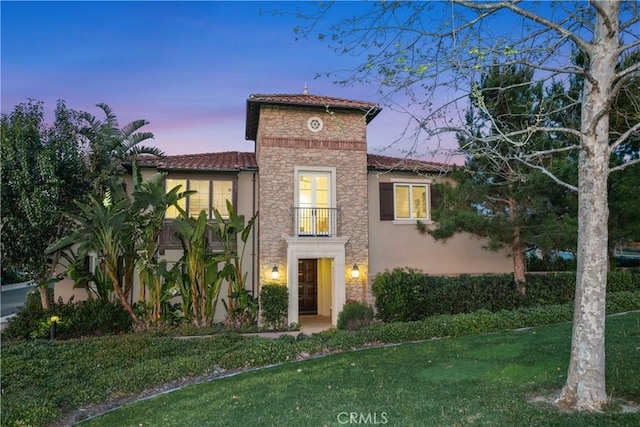 mediterranean / spanish house with a balcony, a tile roof, stone siding, a lawn, and stucco siding