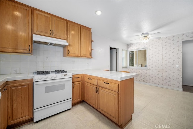 kitchen featuring under cabinet range hood, a peninsula, white range with gas cooktop, tile counters, and wallpapered walls