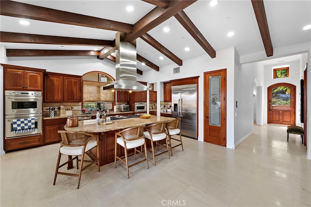 kitchen featuring arched walkways, island exhaust hood, tasteful backsplash, lofted ceiling with beams, and appliances with stainless steel finishes