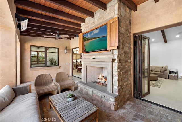 living room featuring wooden ceiling, an outdoor stone fireplace, a ceiling fan, beamed ceiling, and stone tile flooring