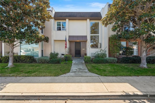 townhome / multi-family property with a shingled roof, a front yard, and stucco siding