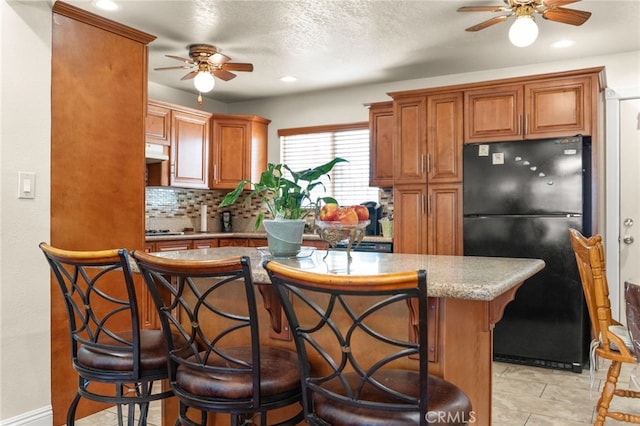 kitchen with brown cabinetry, ventilation hood, backsplash, and freestanding refrigerator