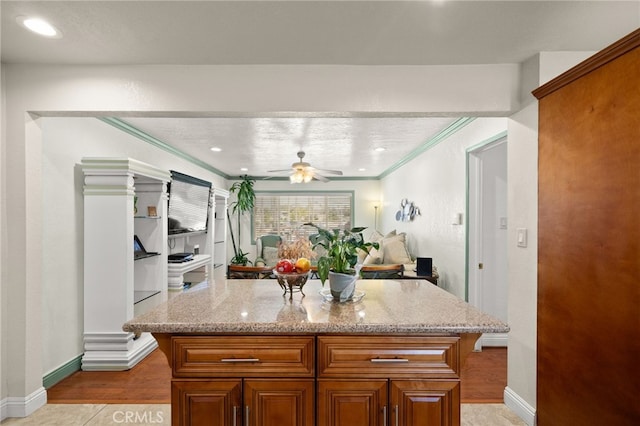 kitchen featuring light stone counters, a center island, brown cabinets, crown molding, and a ceiling fan