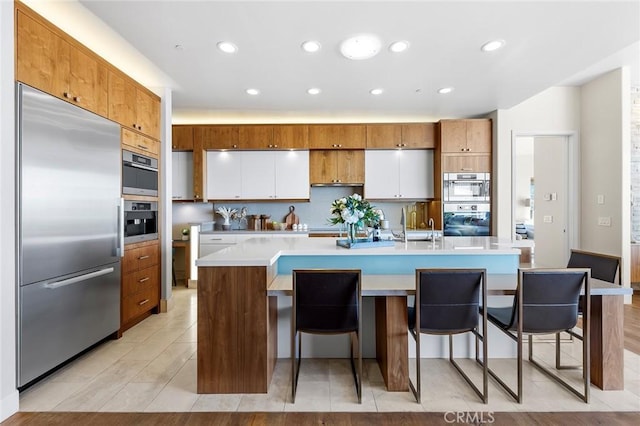 kitchen with white cabinets, an island with sink, brown cabinets, stainless steel appliances, and light countertops