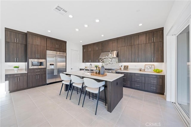 kitchen with light countertops, dark brown cabinetry, built in appliances, under cabinet range hood, and a kitchen breakfast bar