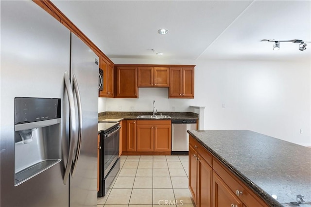 kitchen with appliances with stainless steel finishes, brown cabinetry, light tile patterned flooring, a sink, and dark stone countertops