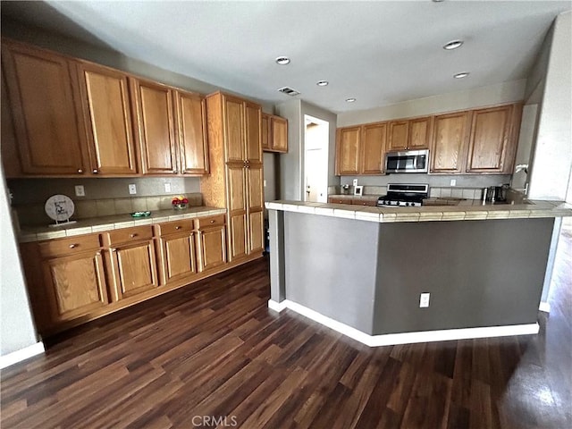 kitchen with tile countertops, brown cabinets, dark wood-style flooring, a peninsula, and stainless steel appliances