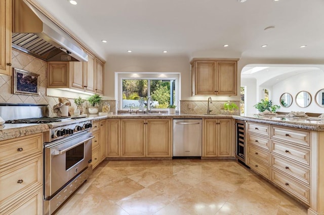 kitchen with appliances with stainless steel finishes, a sink, wall chimney range hood, and light brown cabinetry