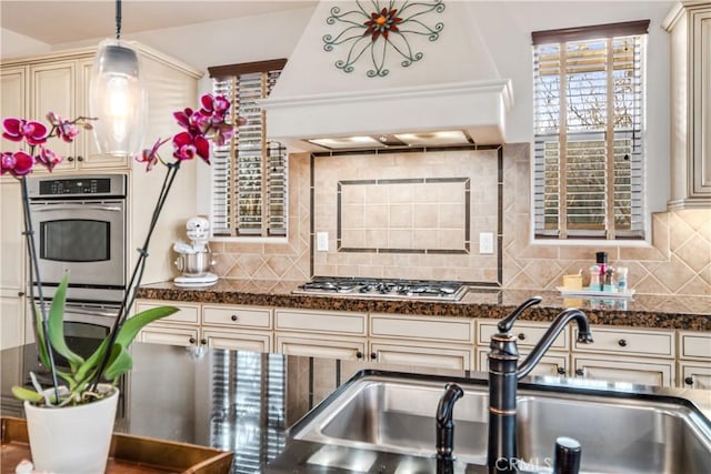 kitchen with cream cabinets, stainless steel appliances, premium range hood, pendant lighting, and a sink