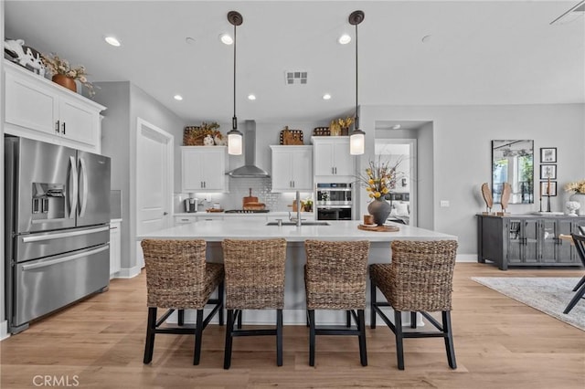 kitchen featuring light countertops, appliances with stainless steel finishes, white cabinets, wall chimney range hood, and an island with sink