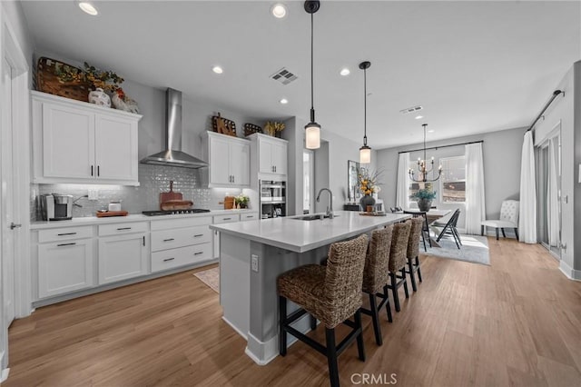 kitchen featuring visible vents, light countertops, white cabinets, wall chimney range hood, and an island with sink