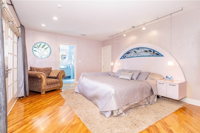 bedroom featuring wood finished floors, visible vents, baseboards, ensuite bath, and track lighting