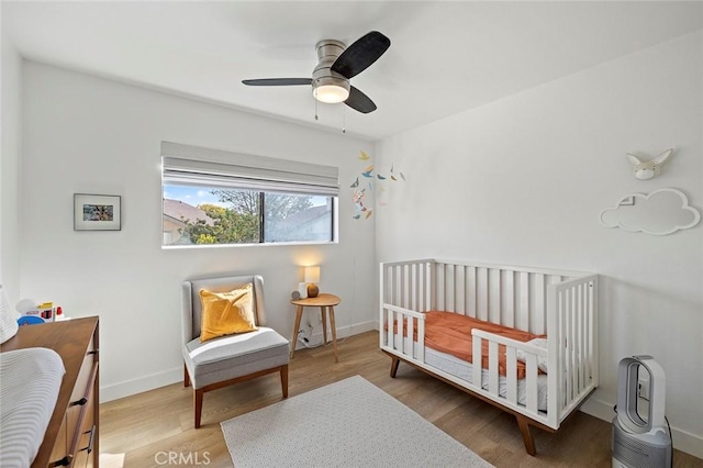 bedroom with a nursery area, ceiling fan, baseboards, and wood finished floors