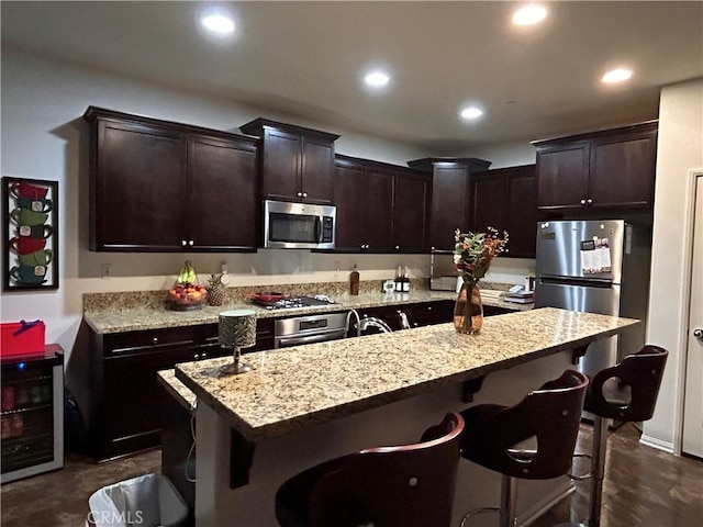 kitchen featuring light stone counters, a center island with sink, stainless steel appliances, dark brown cabinetry, and a kitchen bar