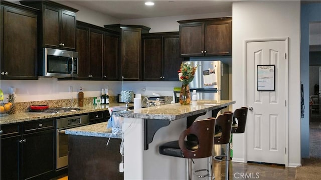 kitchen with light stone counters, a kitchen island with sink, dark brown cabinetry, a breakfast bar, and appliances with stainless steel finishes