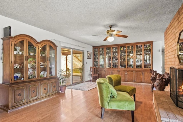 sitting room with light wood-type flooring, a brick fireplace, a textured ceiling, and a ceiling fan