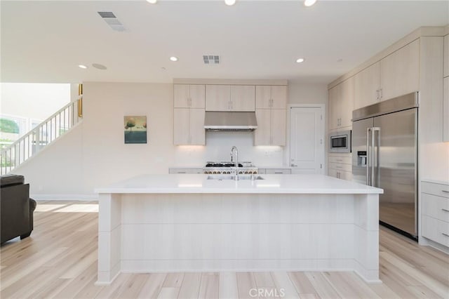 kitchen featuring a center island with sink, visible vents, built in appliances, light countertops, and under cabinet range hood