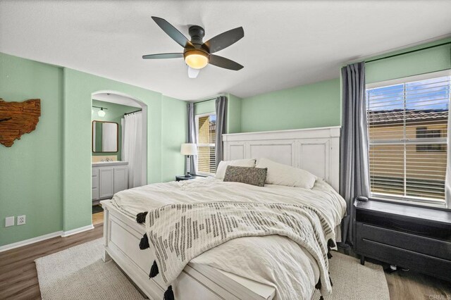 bedroom with arched walkways, connected bathroom, wood finished floors, a ceiling fan, and baseboards