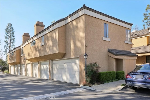 view of side of property featuring stucco siding, a chimney, and community garages