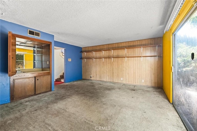 unfurnished room featuring a textured ceiling, wooden walls, carpet floors, visible vents, and stairs