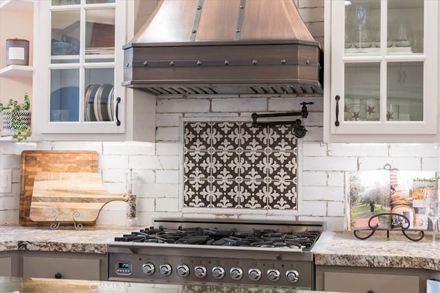 kitchen with light stone counters, backsplash, glass insert cabinets, and custom range hood