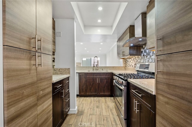 kitchen with a sink, light wood-style floors, wall chimney range hood, decorative backsplash, and gas range