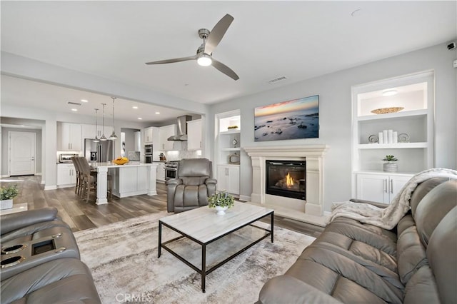 living area with visible vents, a ceiling fan, built in features, light wood-type flooring, and a glass covered fireplace