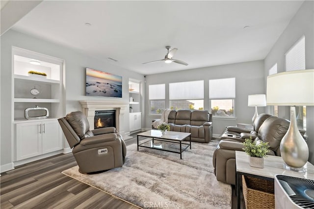 living area with a ceiling fan, a glass covered fireplace, built in features, and wood finished floors